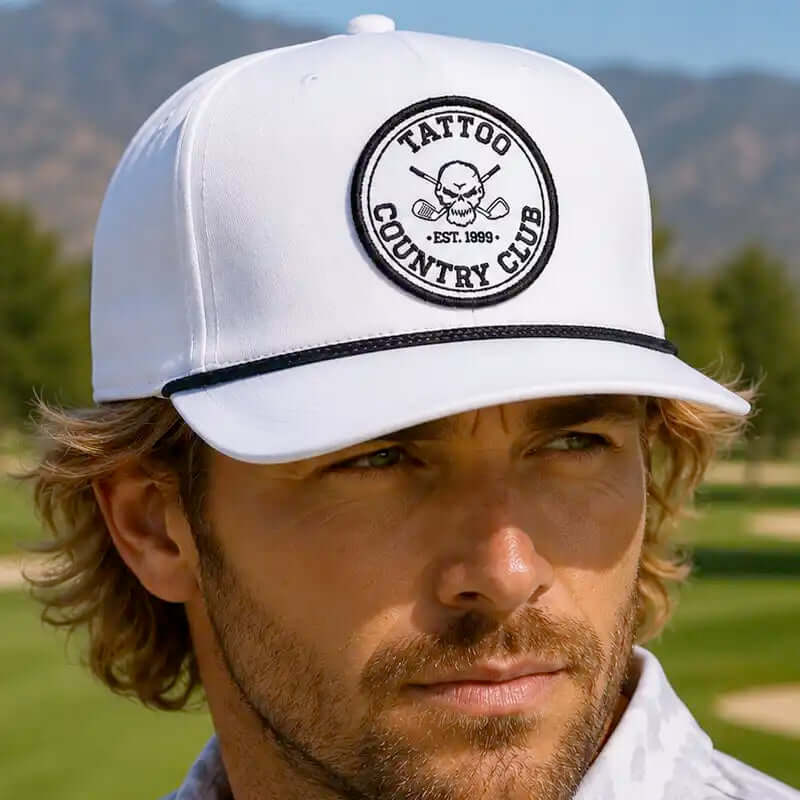 A golfer wearing a white golf hat with a black embroidered circular patch that reads 'TATTOO COUNTRY CLUB EST. 1993' and a black brim stripe, photographed outdoors on a golf course.