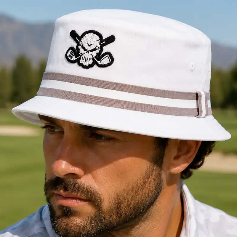 A golfer wearing a white bucket golf hat with a black and white skull and crossed golf clubs patch on the front, and two brown stripes around the crown.