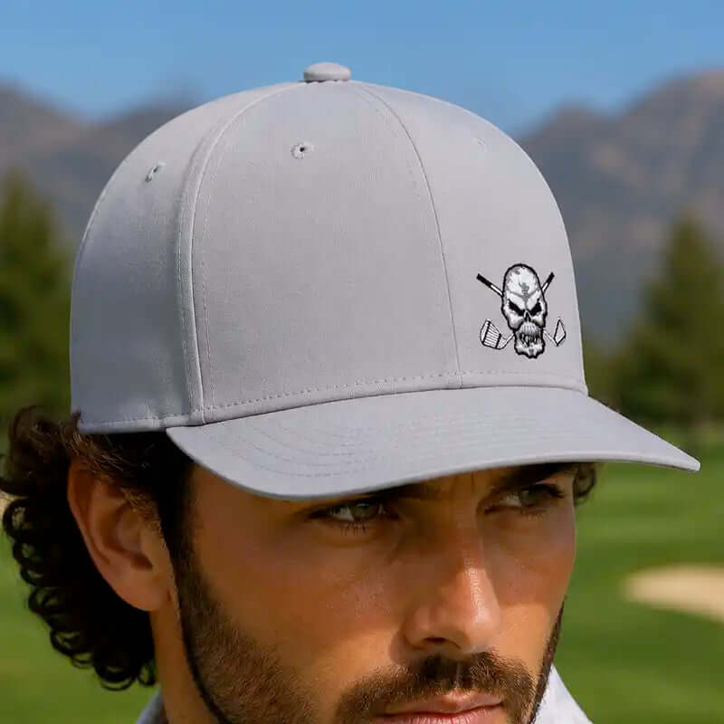 A golfer wearing a light gray baseball cap with a black and white skull-and-crossed-club graphic on the front, photographed outdoors on a golf course with mountains in the background.