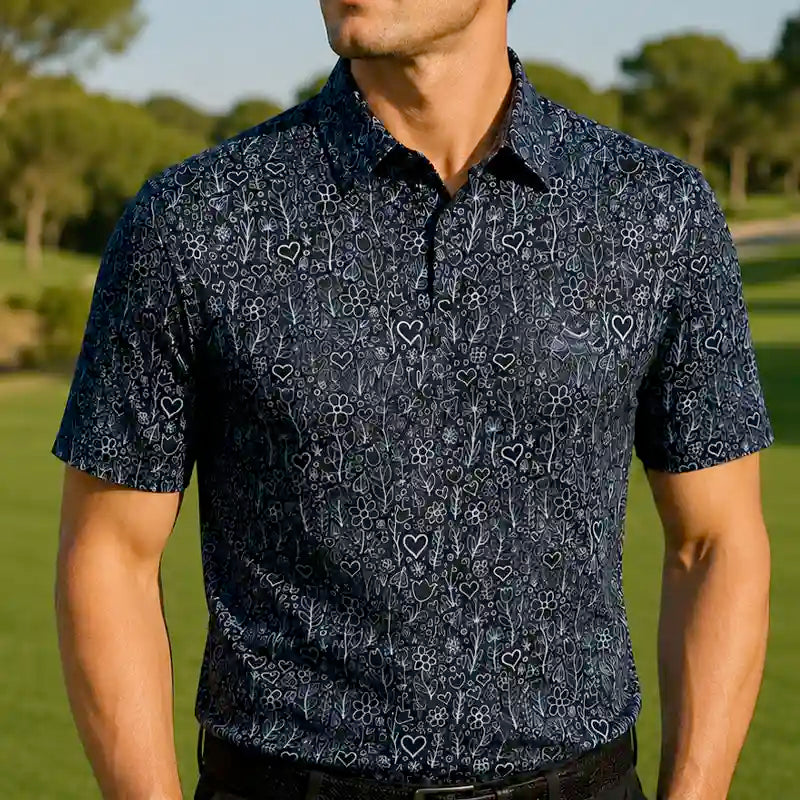 A man wearing a black golf shirt with a dark navy blue and black floral and heart pattern, photographed outdoors on a golf course.