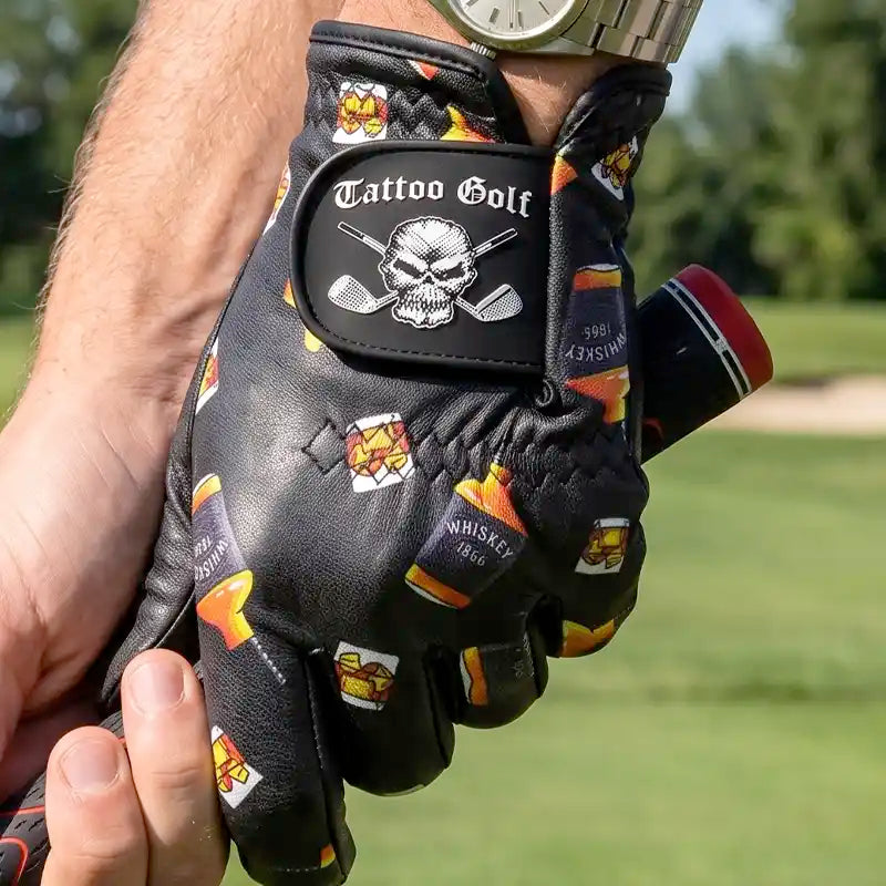 A black leather golf glove with a skull and cross golf clubs patch and whiskey bottle graphics, held by a person's hand on a golf course.