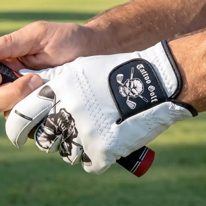 A close-up of a hand wearing a white golf glove with a black patch that reads 'Tattoo Golf' and a skull-and-crossed-golf-clubs logo