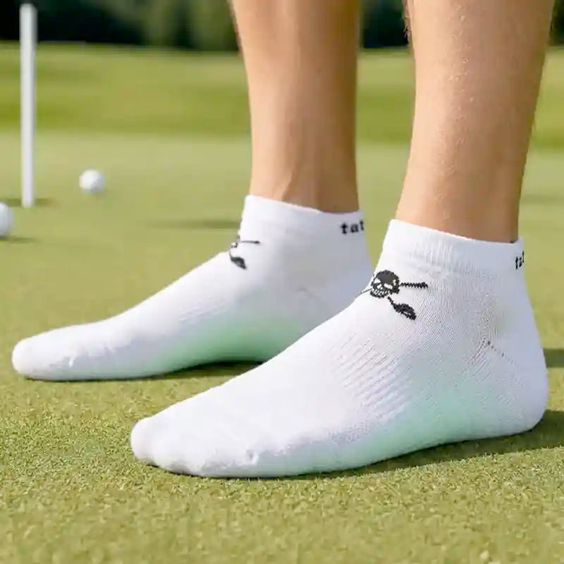 White low-cut athletic socks worn on a person's feet on a green golf course, featuring a small black skull-and-crossclubs graphic
