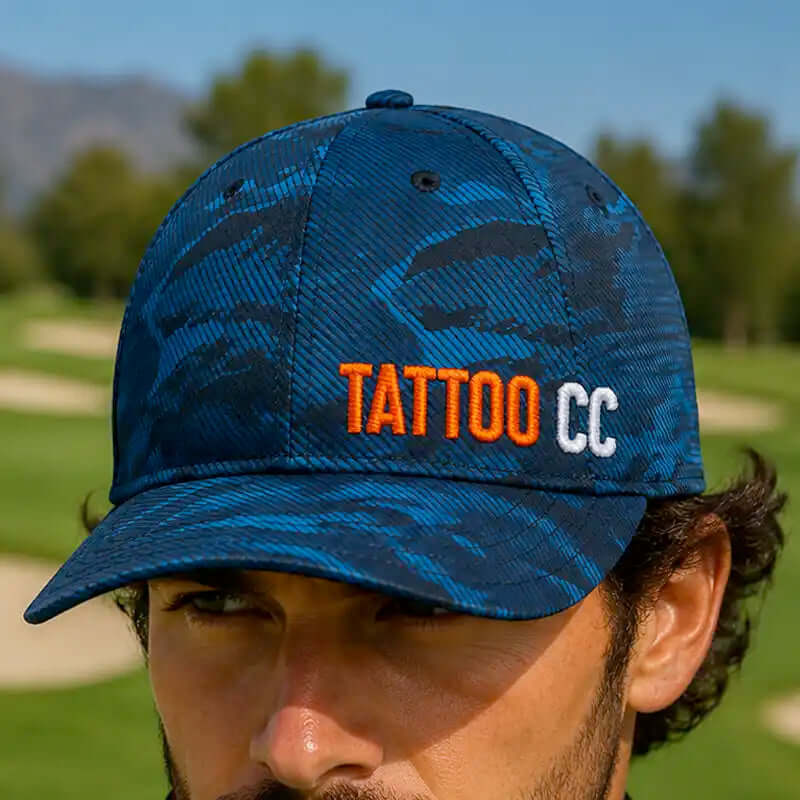 A close-up of a man wearing a blue camouflage golf cap with 'TATTOO CC' embroidered on the front, set against a blurred golf course background.