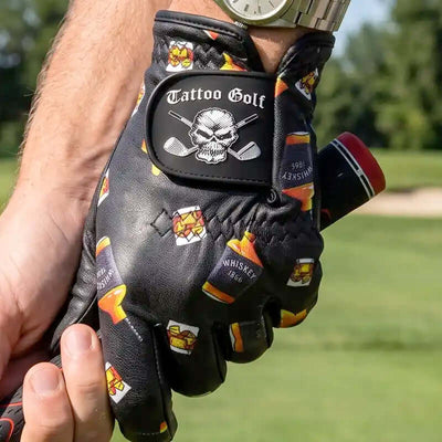 A black leather golf glove with a skull and cross golf clubs patch and whiskey bottle graphics, held by a person's hand on a golf course.