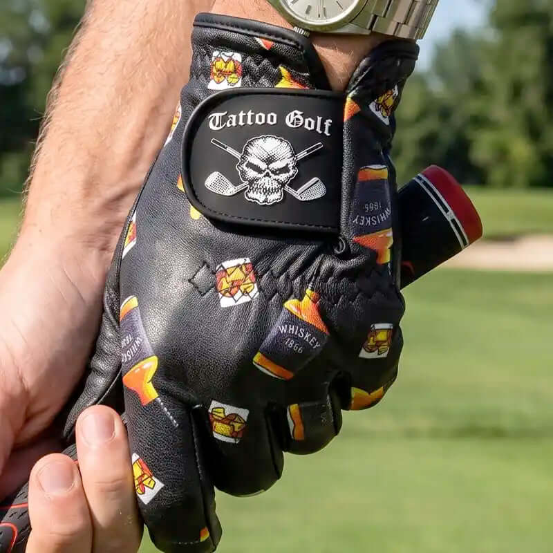 A black leather golf glove with a skull and cross golf clubs patch and whiskey bottle graphics, held by a person's hand on a golf course.
