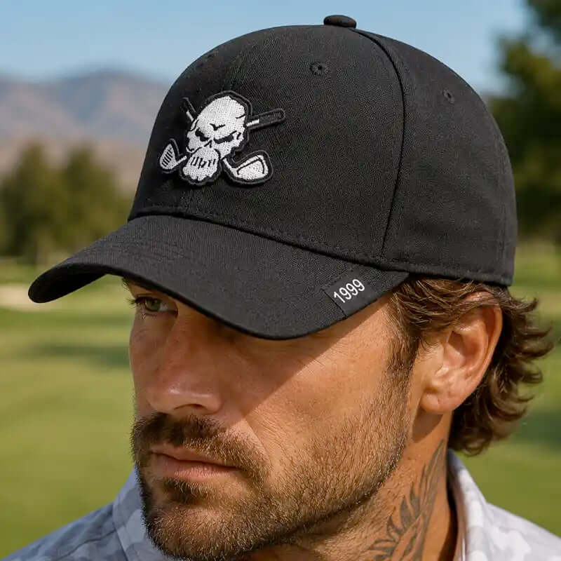 A man wearing a black baseball cap with a white embroidered skull and crossed golf clubs on the front, and a small white '1999' detail on the brim, photographed outdoors on a golf course.