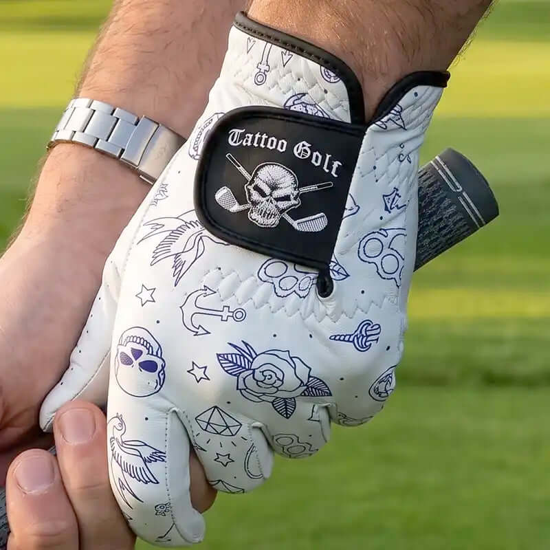 A white golf glove with black and blue tattoo-style graphics, including a skull patch with the words 'Tattoo Golf,' worn on a hand and holding a golf club on a grassy field.