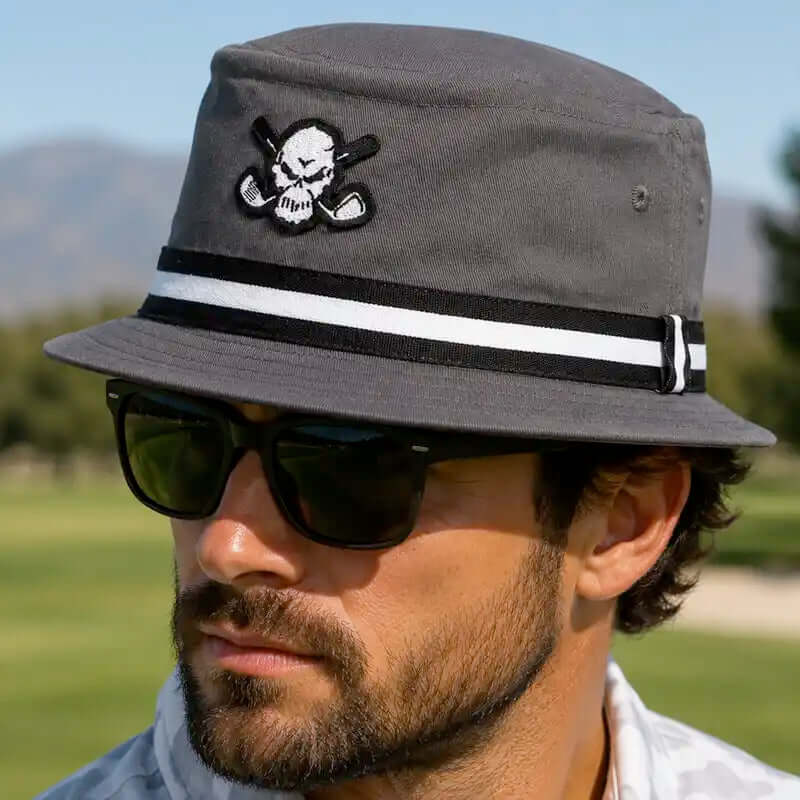 A man wearing a dark gray golf bucket hat with a black and white striped band and a skull patch, along with black sunglasses