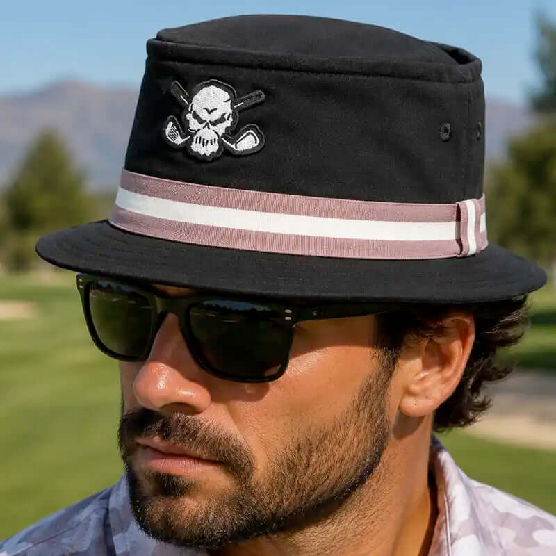 A man wearing a black bucket-style golf hat with a white skull and crossed golf clubs patch on the front, a pink and white striped band, and black sunglasses,