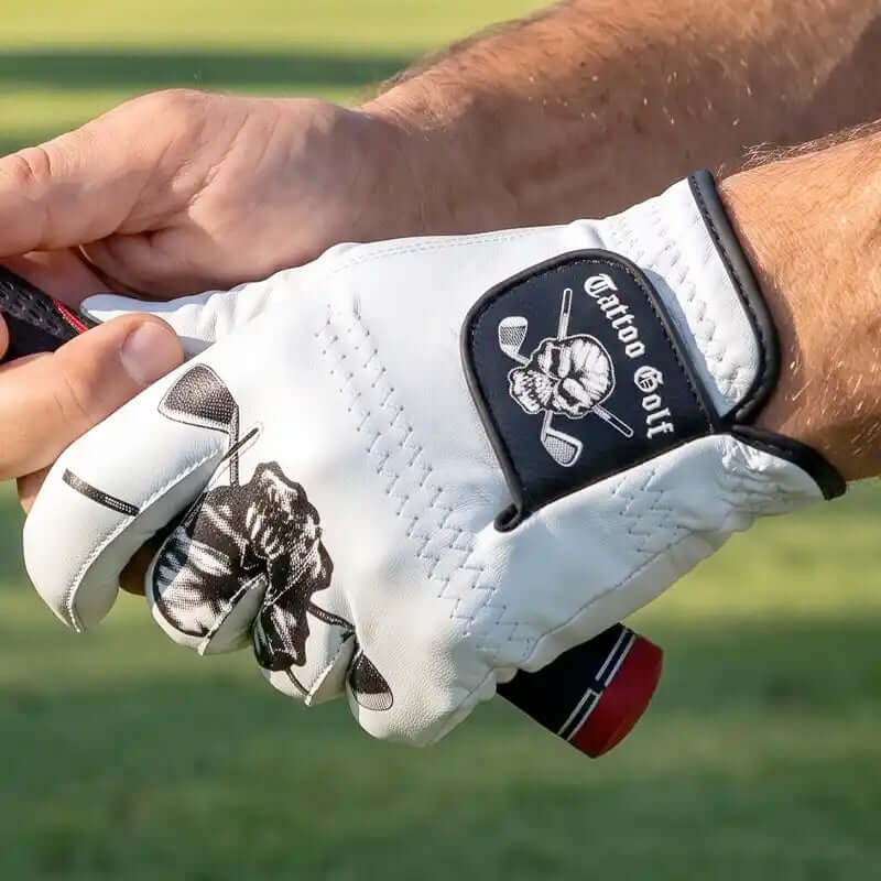 A close-up of a hand wearing a white golf glove with a black patch that reads 'Tattoo Golf' and a skull-and-crossed-golf-clubs logo