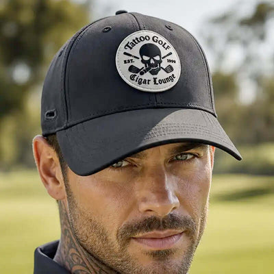 A man wearing a black golf cap with a circular embroidered patch that reads 'Tattoo Golf Est. 2009 Cigar Lounge' featuring a skull and crossed cigars, set against a blurred green golf course background.