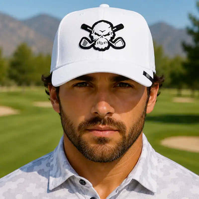 A golfer wearing a white golf cap with a black skull and crossed golf clubs patch, standing on a golf course with mountains in the background.