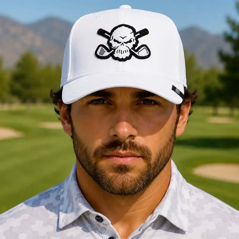 A golfer wearing a white golf cap with a black skull and crossed golf clubs patch, standing on a golf course with mountains in the background.