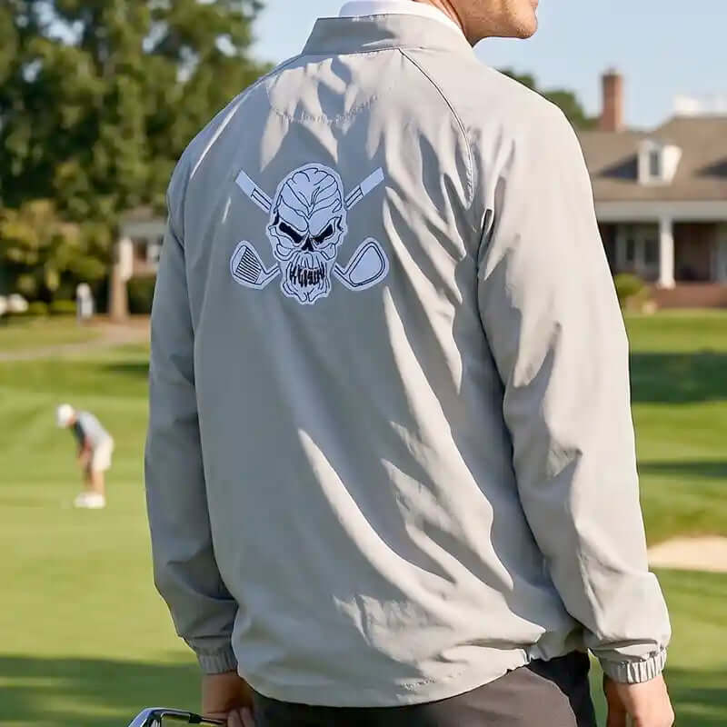 golfer wearing a light gray jacket with a skull and golf clubs design on a golf course.