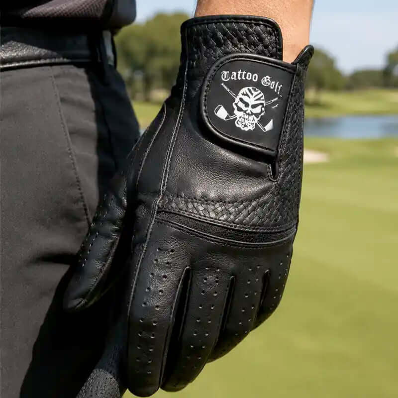 a golfer wearing a black leather golf glove with a white 'Tattoo Golf' skull logo, set against a blurred green golf course background.