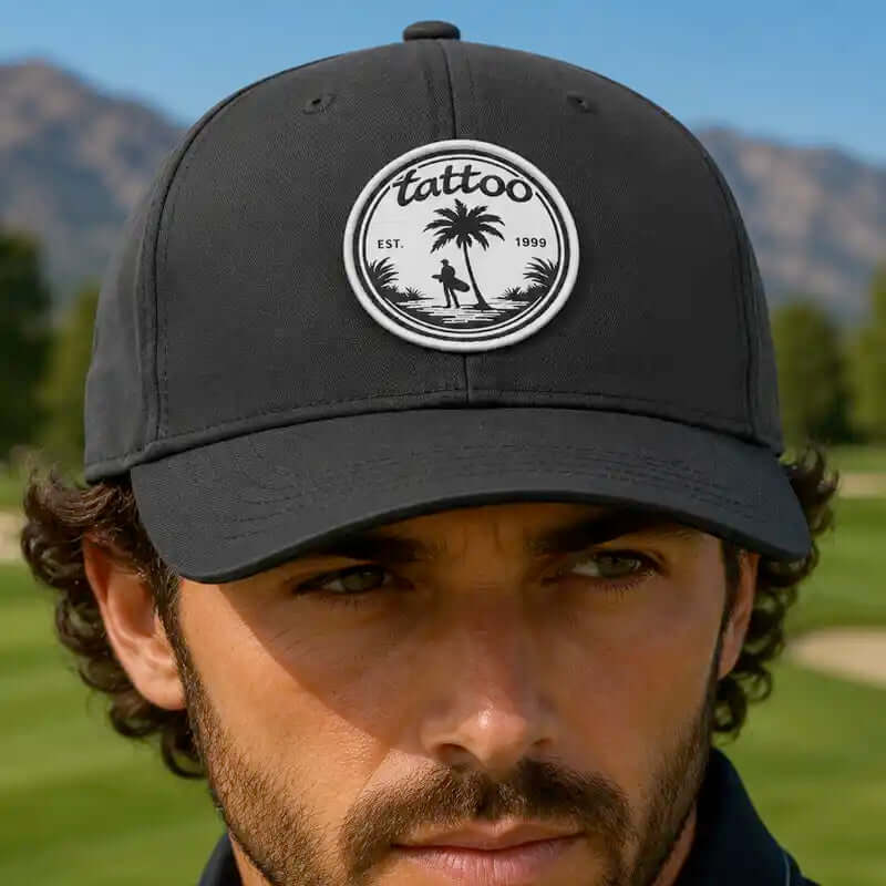 A man wearing a black golf cap with a circular white embroidered patch on the front that reads 'tattoo est. 1999' with a palm tree and figure design