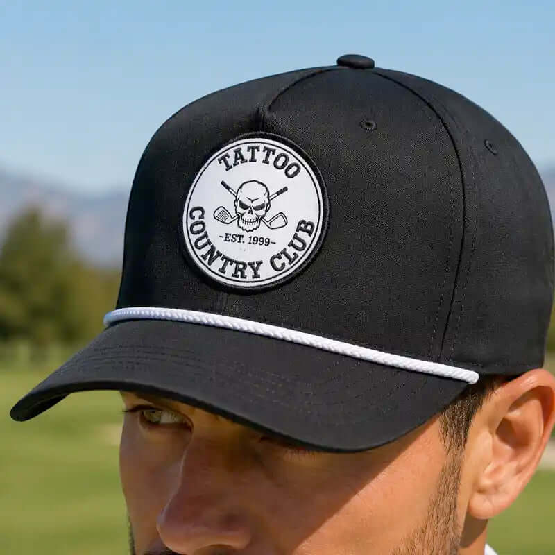 A close-up of a man wearing a black golf cap with a white rope detail and a circular embroidered patch that reads 'TATTOO COUNTRY CLUB EST. 1999' with a skull and crossed clubs