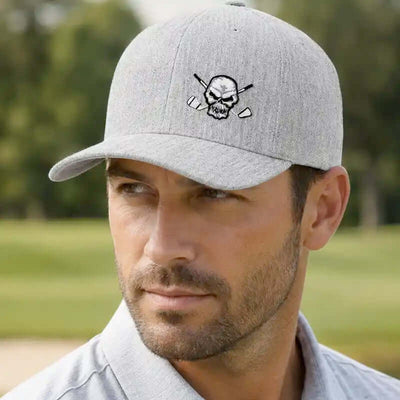 A man wearing a light gray heather golfer cap with an embroidered skull and crossed golf clubs logo on the front, set against a blurred green golf course background.