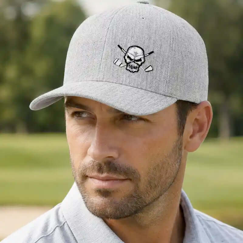 A man wearing a light gray heather golfer cap with an embroidered skull and crossed golf clubs logo on the front, set against a blurred green golf course background.