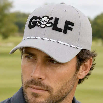 A man wearing a light gray golf cap for men with 'GOLF' embroidered in black across the front, featuring a skull-and-crossbones motif inside the letter O and a braided trim detail on the brim, set against a blurred golf course background.