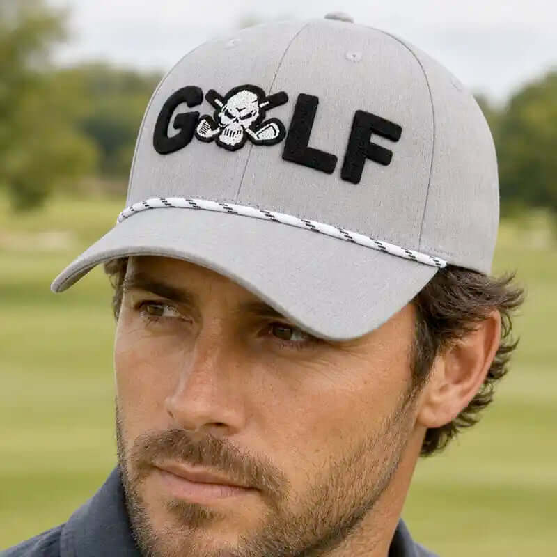 A man wearing a light gray golf cap for men with 'GOLF' embroidered in black across the front, featuring a skull-and-crossbones motif inside the letter O and a braided trim detail on the brim, set against a blurred golf course background.