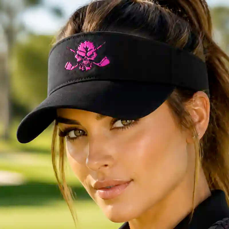 A close-up of a woman wearing a black visor with a pink skull and golf clubs graphic on the front, set against a blurred outdoor background.