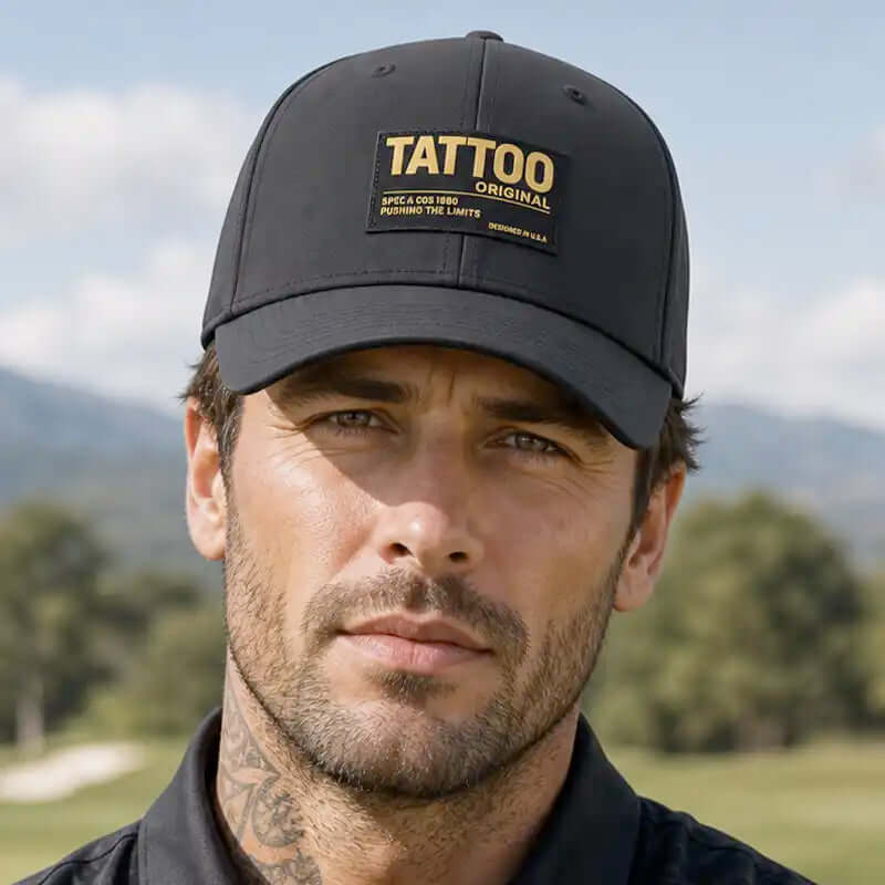A man wearing a black golf hat with a gold and white 'TATTOO ORIGINAL' patch on the front, photographed outdoors with a blurred golf course and mountains in the background.