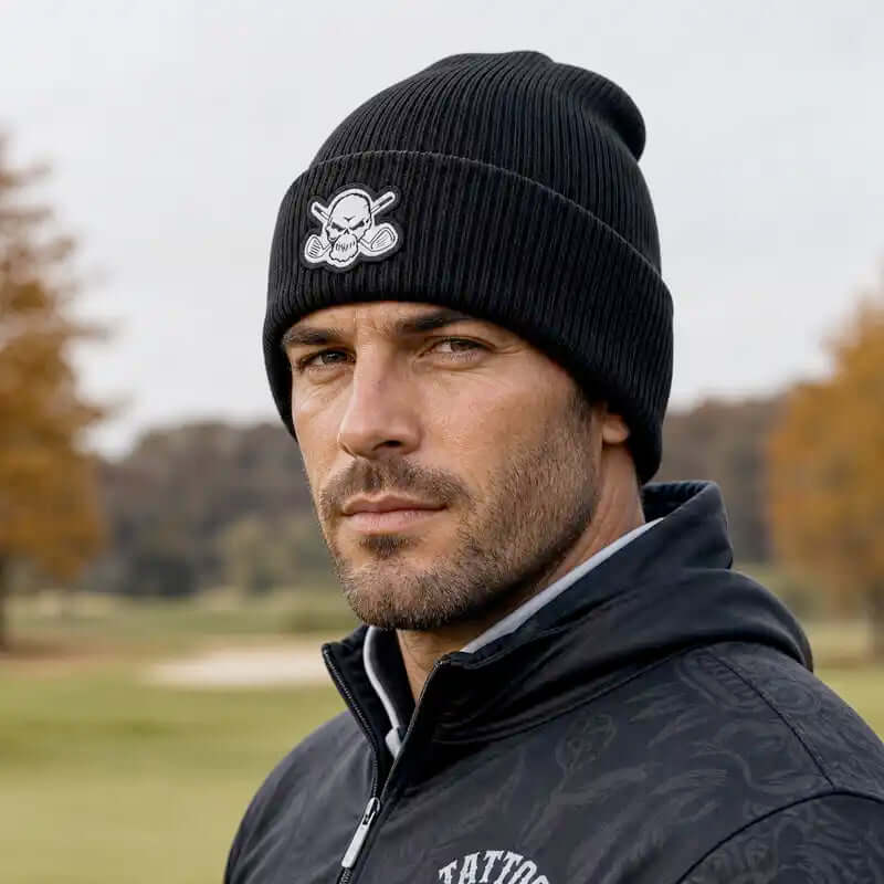 A man wearing a black ribbed knit golf beanie with a white skull and crossbones logo, paired with a dark zip-up hoodie