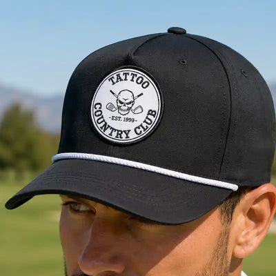 A close-up of a man wearing a black golf cap with a white rope detail and a circular embroidered patch that reads 'TATTOO COUNTRY CLUB EST. 1999' with a skull and crossed clubs
