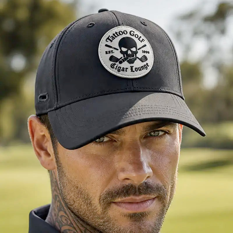 A man wearing a black golf cap with a circular embroidered patch that reads 'Tattoo Golf Est. 2009 Cigar Lounge' featuring a skull and crossed cigars, set against a blurred green golf course background.