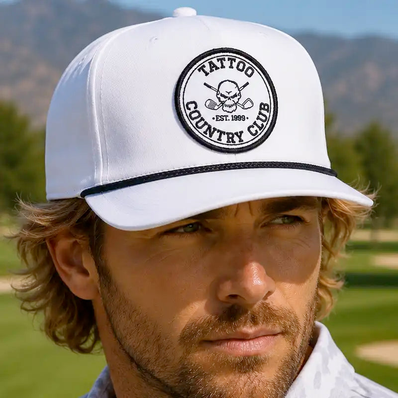 A golfer wearing a white golf hat with a black embroidered circular patch that reads 'TATTOO COUNTRY CLUB EST. 1993' and a black brim stripe, photographed outdoors on a golf course.