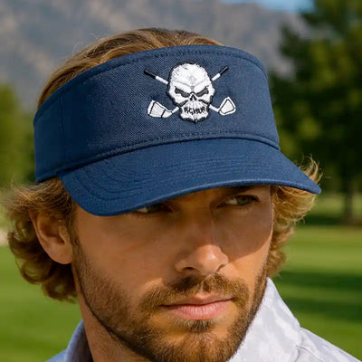 A man wearing a navy blue golf visor hat with a white embroidered skull and crossed golf clubs design on the front, set against a blurred outdoor golf course background.
