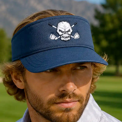 A man wearing a navy blue golf visor hat with a white embroidered skull and crossed golf clubs design on the front, set against a blurred outdoor golf course background.