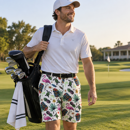 Golfer wearing a white polo and multicolor Tiki golf shorts, carrying a golf bag on a sunlit course.