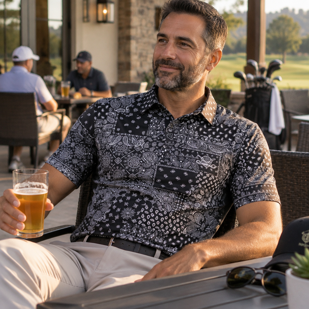 Man relaxing on a patio in a black and grey bandana-print polo, holding a drink.