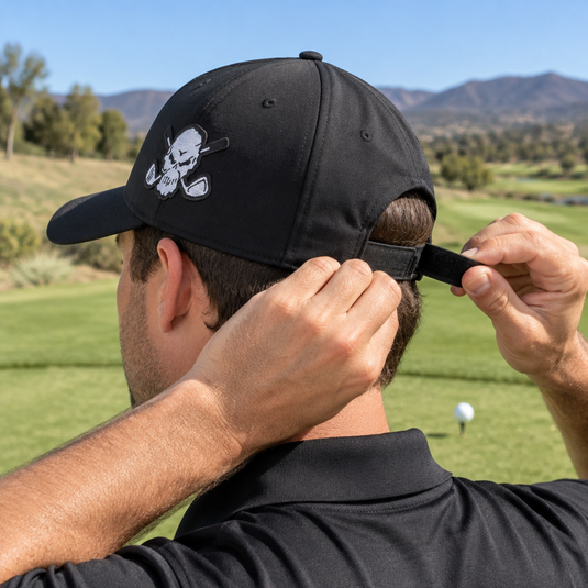 Golfer adjusting the back strap of a black skull-patch cap.