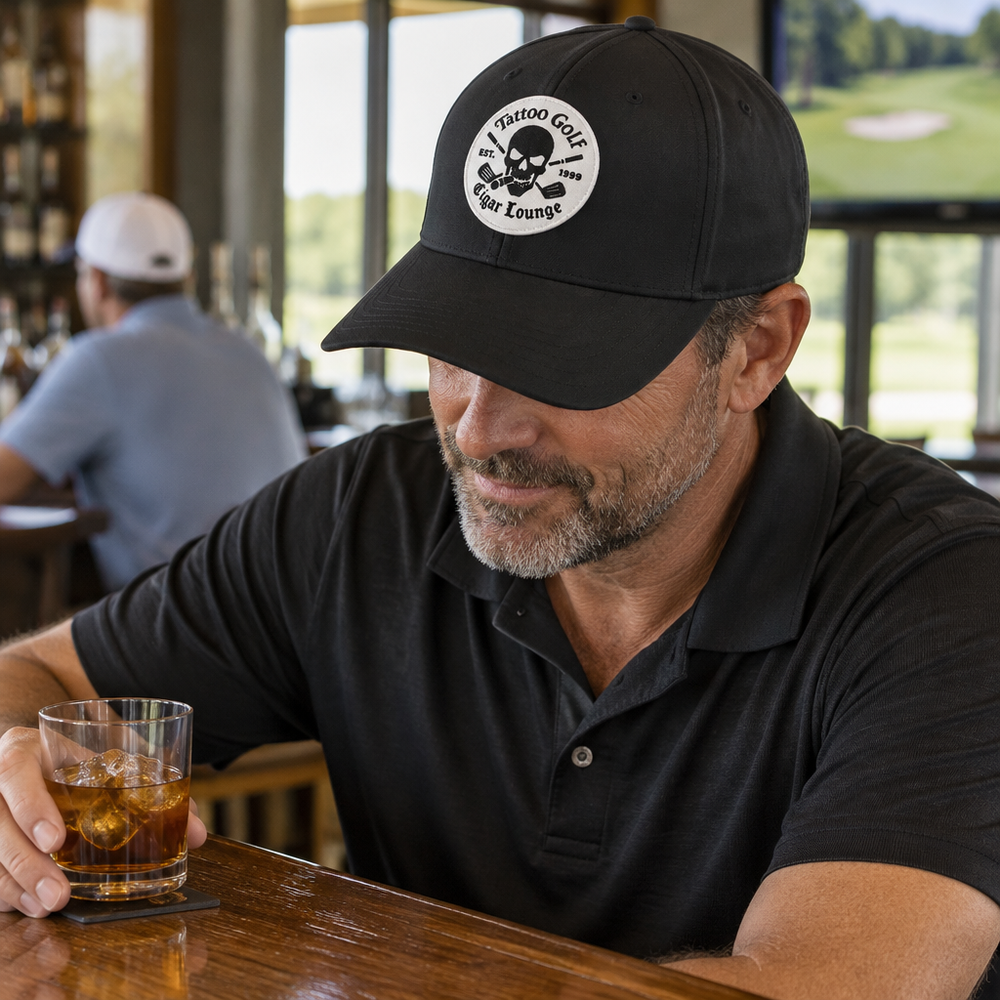 Man seated at a bar wearing a black Cigar Lounge cap and holding a drink.