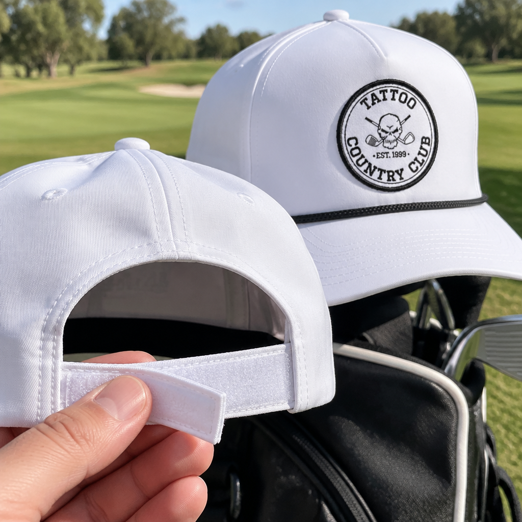 Hand lifting the adjustable back strap of a white cap, with a Tattoo Country Club cap behind it.