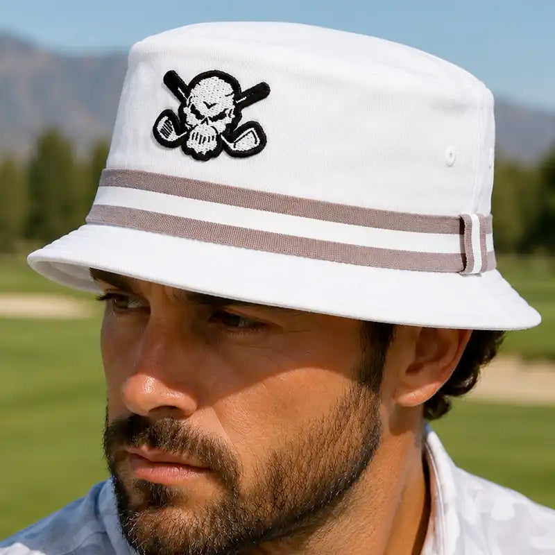 A golfer wearing a white bucket golf hat with a black and white skull and crossed golf clubs patch on the front, and two brown stripes around the crown. 