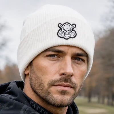 A man wearing a white ribbed knit beanie with a black and white skull-and-crossbones style embroidered patch on the front